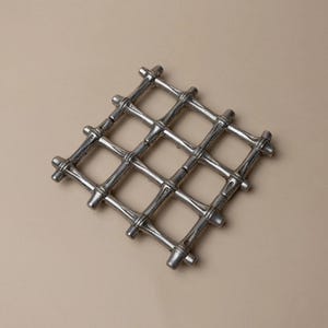May include: A silver-colored, square trivet with a grid pattern. The trivet is made of metal and has a bamboo-like design. The trivet is designed to protect surfaces from hot cookware. The trivet is a decorative kitchen accessory.