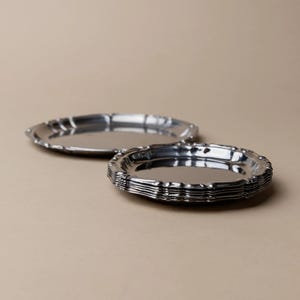 May include: A stack of silver-colored, oval-shaped serving trays with a decorative, scalloped edge. One tray is separate, and the others are stacked. The trays are reflective and sit on a neutral background.
