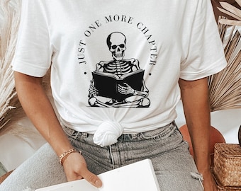 May include: White t-shirt with a black skeleton reading a book. The text on the shirt says "Just One More Chapter".