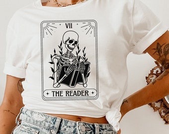 May include: A white t-shirt with a black and white illustration of a tarot card. The card is the VII of Death, also known as The Reader. The card features a skeleton sitting with a book in its lap. The text on the card reads "VII THE READER".