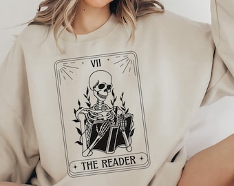 May include: A beige sweatshirt with a black and white illustration of a skeleton reading a book. The illustration is in the style of a tarot card and includes the text "VII THE READER".