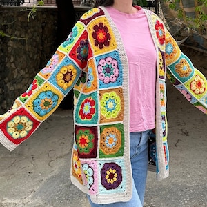 May include: A vibrant, open-front crochet cardigan with square panels displaying floral and geometric patterns in multiple colours. The cardigan has a cream border and is worn over a pink top and blue jeans. The style is casual and handmade.