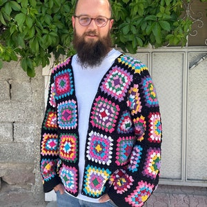 Könnte beinhalten: Ein farbenfroher, offener Häkelcardigan mit einem Patchwork-Design aus Quadraten in verschiedenen Farben, darunter Rosa, Blau, Gelb und Grün, vor einem schwarzen Hintergrund. Der Cardigan wird über einem weißen Hemd und einer blauen Jeans getragen.
