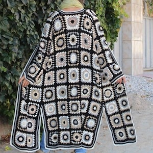 Op de afbeelding: Lange, open gehaakte vest met een granny square patroon. Het kleurenpalet omvat zwart, wit en verschillende tinten beige en bruin. Het vest heeft lange mouwen en een split aan de onderkant. De persoon draagt een groene sjaal.