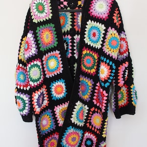 Può includere: Un cardigan nero aperto sul davanti con un colorato design a quadretti granny. Il maglione all'uncinetto presenta quadrati in vari colori, tra cui rosa, giallo, verde e blu. Il capo a maniche lunghe è esposto su una gruccia nera.