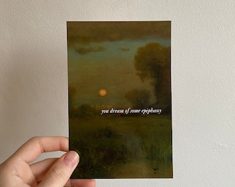 May include: A rectangular art print depicting a landscape scene with a sunset, trees, and grassy areas. The text "you dream of some epiphany" is printed in white. The print is held by a hand.