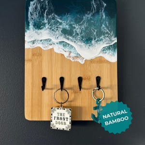 May include: A key holder made of natural bamboo with a resin ocean wave design. The key holder has four hooks and a "The Front Door" keychain attached to one of the hooks. The key holder has a teal "Natural Bamboo" sticker on the bottom right corner.