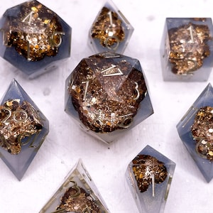 May include: A set of seven polyhedral dice. Each die is clear with a smoky blue hue, encasing gold flakes and glitter. The numbers are metallic gold. The dice are arranged on a white surface.