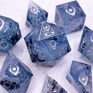 Könnte beinhalten: Satz von sieben blauen und silbernen Würfeln mit einem wirbelnden Muster und einem Augensymbol auf jeder Seite. Die Würfel sind aus klarem Harz mit eingebettetem silbernen Glitter.