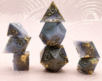 May include: A set of six polyhedral dice with a dark blue and grey color scheme. The dice are decorated with gold leaf and have a variety of numbers and symbols on their faces.