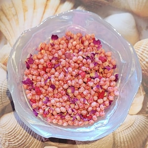 May include: A clear, iridescent glass bowl filled with small, round, pink beads and scattered dried rose petals. The bowl sits on a background of seashells. The beads have a speckled appearance, and the petals are a deep red.