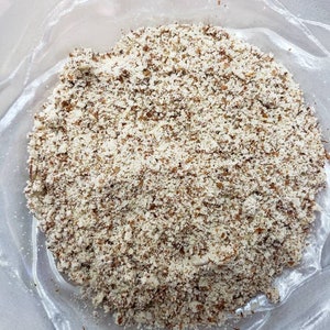 May include: A close-up shot of a clear plastic bag filled with coarsely ground almonds. The almonds are a mix of light beige and brown, creating a textured appearance. The bag is slightly transparent, allowing the contents to be visible.
