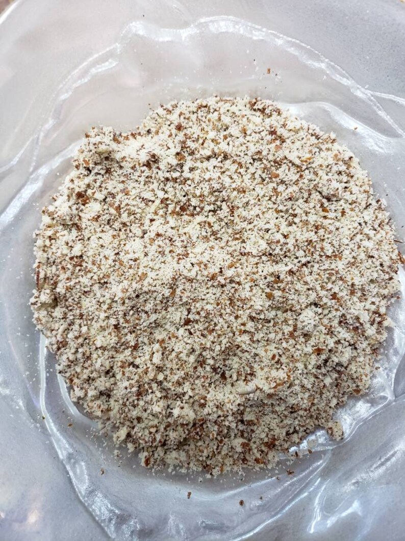 May include: A close-up shot of a clear plastic bag filled with coarsely ground almonds. The almonds are a mix of light beige and brown, creating a textured appearance. The bag is slightly transparent, allowing the contents to be visible.