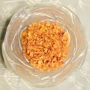 May include: A clear glass bowl filled with diced, orange-colored food. The bowl has a wavy, organic shape. The food appears to be small, uniform pieces, possibly dried fruit or vegetables.