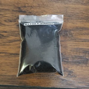 May include: A clear plastic bag filled with dark, granular material, labeled "Worm Castings" in white text. The bag is sealed and rests on a wooden surface with a visible wood grain.