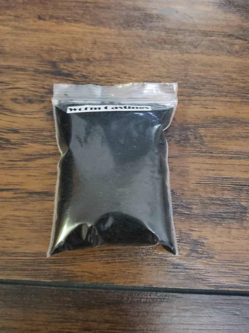 May include: A clear plastic bag filled with dark, granular material, labeled "Worm Castings" in white text. The bag is sealed and rests on a wooden surface with a visible wood grain.