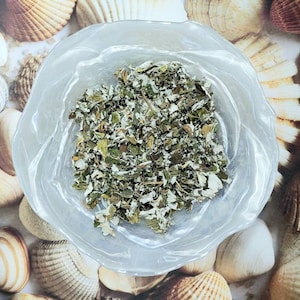 May include: A clear glass bowl filled with dried green and white herbs. The bowl is sitting on a surface with a pattern of seashells.