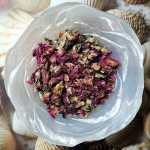 May include: A white bowl filled with dried rose petals. The petals are a mix of pink and red, and some are still attached to the green stems.