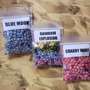 May include: Three small clear plastic bags filled with colorful, speckled beads. Each bag has a white label with text: "BLUE MOON", "RAINBOW EXPLOSION", and "CRABBY MARY". The beads are blue, rainbow-colored, and pink, respectively. The background is a sandy surface.