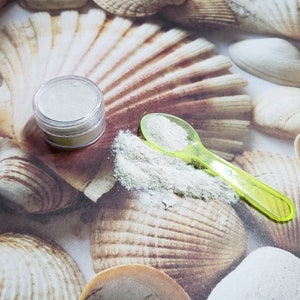 May include: A small clear jar filled with white powder sits on a surface of seashells. A green plastic spoon with white powder in it is next to the jar.