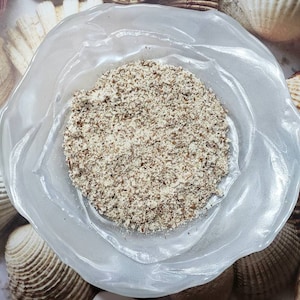 May include: A clear, petal-shaped glass bowl filled with finely ground, light brown food product. The bowl sits on a surface with seashell imagery. The food product appears to be a nut or seed-based ingredient, possibly for culinary use.