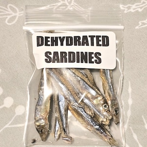 May include: A clear plastic bag containing dehydrated sardines. The bag has a white label with the words "DEHYDRATED SARDINES" in bold, black capital letters. The sardines are small, silver, and dried.