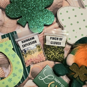 May include: A St. Patrick's Day themed image with a green glitter shamrock, a wooden horseshoe, and a gnome. Two small bags are labeled "Rainbow Explosion" and "Pinch of Clover".