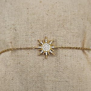 Peut inclure: Un bracelet en chaîne dorée avec un pendentif soleil incrusté de strass transparents. Le bracelet est présenté sur une surface en tissu beige.