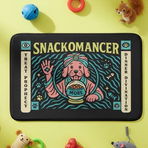 A rectangular pet feeding mat featuring a retro mystical graphic titled SNACKOMANCER in beige arched text. The center illustration depicts a pink dog wearing a teal turban, waving one paw and gazing into a crystal ball that is actually a bowl of food labeled MORE. The background is black with teal wavy lines and floating fish bones. Side panels in beige and teal feature paw prints and text reading TREAT PROPHECY and DINNER DIVINATION. The mat is made of neoprene with a non-slip backing.