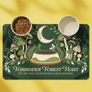 May include: A green pet food mat with a whimsical "Forbidden Forest Feast" design, featuring mushrooms, a crescent moon, and the text "Do Not Feed After Midnight (Just Kidding)." Two white ceramic bowls sit on the mat, one filled with brown pet food.