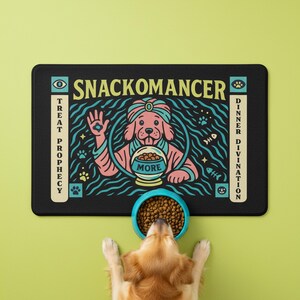 A rectangular pet feeding mat featuring a retro mystical graphic titled SNACKOMANCER in beige arched text. The center illustration depicts a pink dog wearing a teal turban, waving one paw and gazing into a crystal ball that is actually a bowl of food labeled MORE. The background is black with teal wavy lines and floating fish bones. Side panels in beige and teal feature paw prints and text reading TREAT PROPHECY and DINNER DIVINATION. The mat is made of neoprene with a non-slip backing.