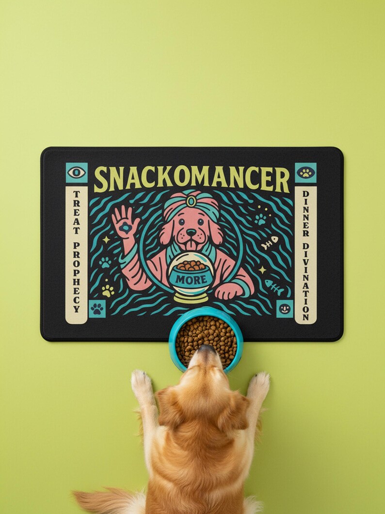 A rectangular pet feeding mat featuring a retro mystical graphic titled SNACKOMANCER in beige arched text. The center illustration depicts a pink dog wearing a teal turban, waving one paw and gazing into a crystal ball that is actually a bowl of food labeled MORE. The background is black with teal wavy lines and floating fish bones. Side panels in beige and teal feature paw prints and text reading TREAT PROPHECY and DINNER DIVINATION. The mat is made of neoprene with a non-slip backing.