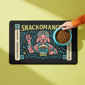 A rectangular pet feeding mat featuring a retro mystical graphic titled SNACKOMANCER in beige arched text. The center illustration depicts a pink dog wearing a teal turban, waving one paw and gazing into a crystal ball that is actually a bowl of food labeled MORE. The background is black with teal wavy lines and floating fish bones. Side panels in beige and teal feature paw prints and text reading TREAT PROPHECY and DINNER DIVINATION. The mat is made of neoprene with a non-slip backing.