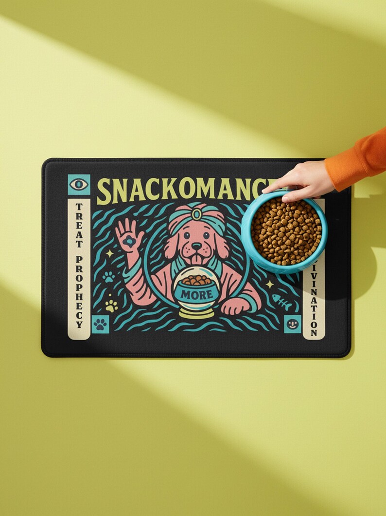A rectangular pet feeding mat featuring a retro mystical graphic titled SNACKOMANCER in beige arched text. The center illustration depicts a pink dog wearing a teal turban, waving one paw and gazing into a crystal ball that is actually a bowl of food labeled MORE. The background is black with teal wavy lines and floating fish bones. Side panels in beige and teal feature paw prints and text reading TREAT PROPHECY and DINNER DIVINATION. The mat is made of neoprene with a non-slip backing.