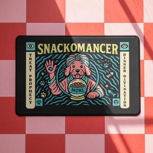 A rectangular pet feeding mat featuring a retro mystical graphic titled SNACKOMANCER in beige arched text. The center illustration depicts a pink dog wearing a teal turban, waving one paw and gazing into a crystal ball that is actually a bowl of food labeled MORE. The background is black with teal wavy lines and floating fish bones. Side panels in beige and teal feature paw prints and text reading TREAT PROPHECY and DINNER DIVINATION. The mat is made of neoprene with a non-slip backing.