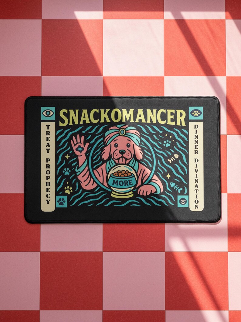 A rectangular pet feeding mat featuring a retro mystical graphic titled SNACKOMANCER in beige arched text. The center illustration depicts a pink dog wearing a teal turban, waving one paw and gazing into a crystal ball that is actually a bowl of food labeled MORE. The background is black with teal wavy lines and floating fish bones. Side panels in beige and teal feature paw prints and text reading TREAT PROPHECY and DINNER DIVINATION. The mat is made of neoprene with a non-slip backing.