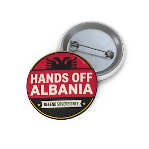 Op de afbeelding: Een ronde button met een speld aan de achterzijde, voorzien van een rode achtergrond met een zwart tweekoppig adelaarsymbool bovenaan. De tekst HANDS OFF ALBANIA staat in grote witte letters, met daaronder een zwarte banner met DEFEND SOVEREIGNTY.
