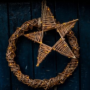 May include: A woven willow wreath with a star-shaped centre, both decorated with warm white fairy lights. The wreath is hanging on a dark blue wooden door.