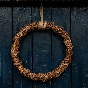May include: A circular woven wreath with warm, glowing lights hangs against a dark blue wooden door. The wreath is made of intertwined brown branches, creating a rustic and natural aesthetic. The lights add a touch of warmth.