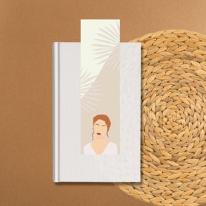 May include: A white book with a bookmark featuring a woman with red hair and a white dress against a background of beige and white palm leaves.