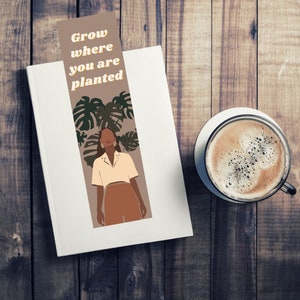 May include: A brown bookmark with the text "Grow where you are planted" in yellow letters. A woman with brown skin and long brown hair is standing in front of a green leafy plant. The bookmark is on a white surface.