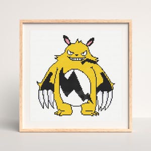 May include: Framed cross-stitch art featuring a yellow and black monster with sharp claws and a menacing expression. The artwork is set against a white background and framed in a light wood frame. The monster has a white belly and a black zig-zag pattern.