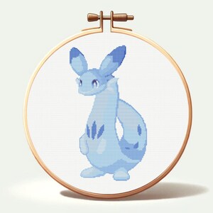 May include: A cross-stitch embroidery of a light blue creature with large ears, a long neck, and a curved tail. The design is set in a wooden embroidery hoop. The creature has darker blue markings and is set against a white background.