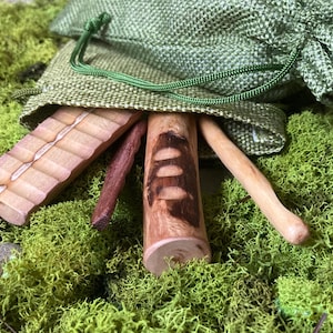 May include: A collection of wooden tools, including a notched block, a dark brown stick, and a cylindrical piece with cut-out designs, partially emerging from a green drawstring bag. The items rest on a bed of green moss.