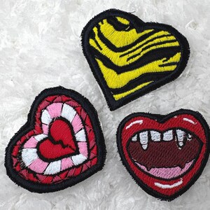 May include: Three heart-shaped embroidered patches. One patch features a yellow and black tiger stripe pattern. Another patch displays a red heart with pink and white concentric borders. The third patch shows a red mouth with white fangs.