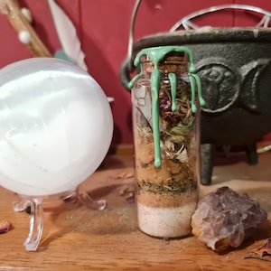 Könnte beinhalten: Eine klare Glasflasche gefüllt mit Schichten aus Kräutern, Sand und getrockneten Blumen, verschlossen mit einem Korken und grünem Wachs. Eine große, klare Kristallkugel steht links auf einem Ständer. Ein schwarzer Kessel ist im Hintergrund.
