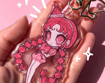 Peut inclure: Un porte-clés en acrylique rose représentant un personnage de dessin animé aux cheveux roses, un chapeau de fête et des yeux en forme de cœur. Le porte-clés est attaché à un fermoir doré en forme de nœud. Le fond est rose uni.