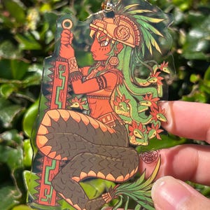 May include: A clear acrylic charm featuring a stylized illustration of a figure with a snake-like lower body, green hair with small snake heads, and an orange headdress. The figure holds a long, rectangular orange and green sword-like object.