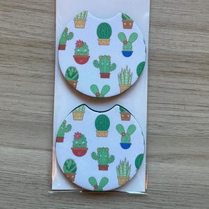 May include: Two car coasters with a white background and a pattern of various cacti in pots. The coasters are packaged in a clear plastic sleeve. The packaging has the words "Car Coasters" and "Handmade with love".