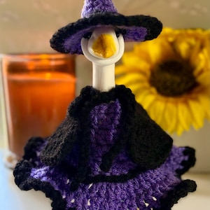 Pu&ograve; includere: Una statuetta di oca in ceramica bianca che indossa un completo lavorato all'uncinetto a mano in viola e nero. L'insieme include un cappello a punta, una piccola mantella e una gonna a balze, tutto con una trama in lana e bordi scuri.
